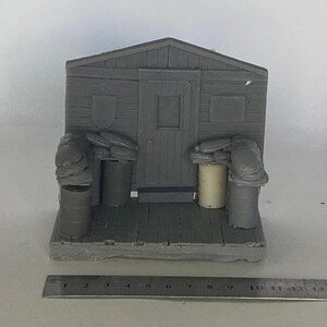 May include: A grey miniature model of a wooden shed with a central door, flanked by stacks of sandbags and cylindrical barrels on a platform base. A metal ruler is positioned in front, showing a width of approximately 13.3 cm for the base.