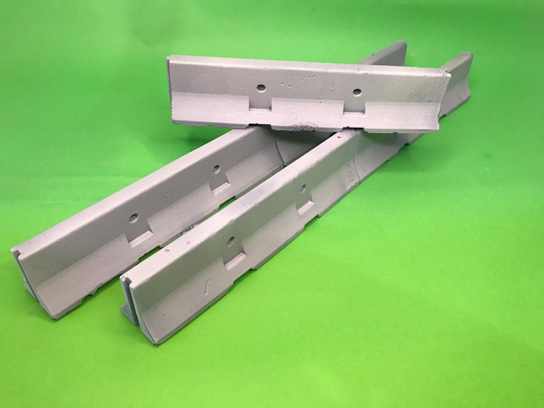 May include: Three light gray concrete jersey barriers arranged in a cluster. Each barrier features a sloped profile, a flat top, and two circular holes along the upper section, mimicking the design of full-scale highway traffic dividers.