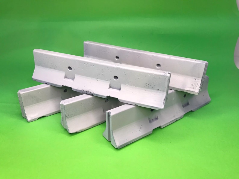 May include: A set of three light gray concrete traffic barriers, each featuring a distinct L-shaped profile with a wide base and a vertical wall. The barriers have small circular holes along the top edge and a textured, industrial surface finish.