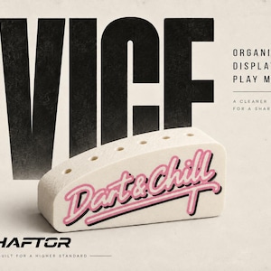 May include: A white, wedge-shaped dart holder with five circular holes along the top edge. The front features the pink and black text 'Dart & Chill'. Large black text reads 'VICE' above, with 'ORGANIZE. DISPLAY. PLAY MORE.' and 'SHAFTOR' below.