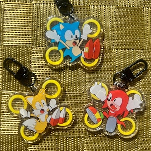 May include: Three clear acrylic keychains featuring Sonic the Hedgehog, Tails, and Knuckles. Each character is depicted in a dynamic pose, surrounded by yellow rings. The keychains have black metal clasps.