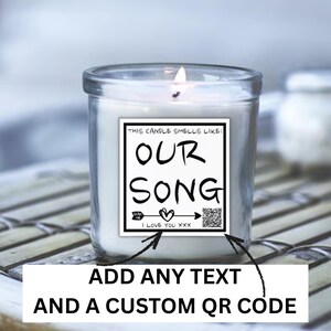 May include: A lit white candle in a clear glass jar. The candle has a white label with the text "THIS CANDLE SMELLS LIKE: OUR SONG" and a QR code. The bottom of the label reads "I LOVE YOU XXX".