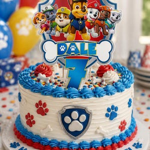 Op de afbeelding: Een ronde taart met wit glazuur, versierd met blauwe en rode randen, pootafdrukken en een centraal schildembleem. Een taarttopper toont de Paw Patrol-figuren, de naam DALE en het cijfer 7. Op de onderkant staat de tekst HAPPY BIRTHDAY DALE.