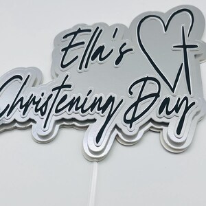Può includere: Un topper per torta a strati argento e nero con il testo Ella's Christening Day in carattere corsivo. Il design include una forma a cuore con un simbolo di croce integrato sul lato destro, il tutto montato su un sottile bastoncino bianco.