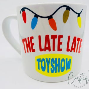 May include: White ceramic mug with a handle, featuring the text "THE LATE LATE TOYSHOW" in red and blue, with a string of colourful lights above. The mug has a glossy finish.