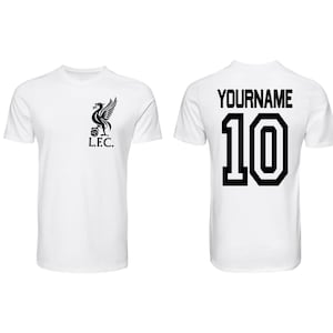 May include: White t-shirt featuring the Liverpool Football Club emblem on the front and a personalized name and number 10 on the back. The emblem is a black bird with the letters L.F.C. below it. The text "YOURNAME" is above the number.