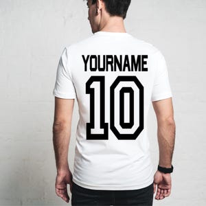 May include: White t-shirt with the name "YOURNAME" and the number "10" printed in black on the back. The shirt is a classic crew neck style, perfect for sports or casual wear. The black text provides a strong contrast against the white fabric.