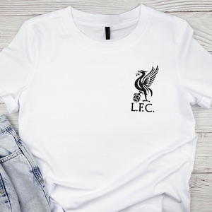 May include: White t-shirt with a black graphic of a bird with outstretched wings, a ball, and the letters "L.F.C." The shirt is made of a soft material and has short sleeves. The shirt is folded on a light wood surface.