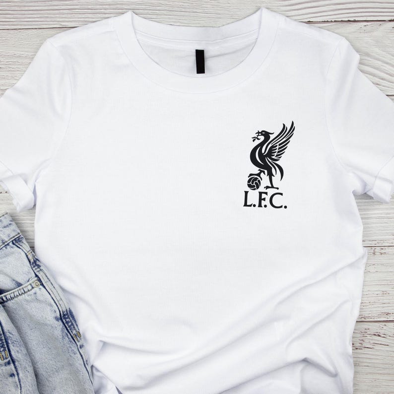May include: White t-shirt with a black graphic of a bird with outstretched wings, a ball, and the letters "L.F.C." The shirt is made of a soft material and has short sleeves. The shirt is folded on a light wood surface.