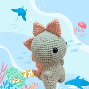 The dimensions of the green and orange knitted amigurumi dinosaur are 13 cm high and 7 cm wide.