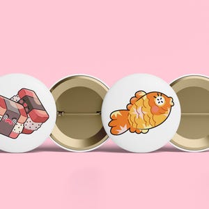 May include: Two circular pin-back buttons with white backgrounds. One button features a cartoon illustration of two pieces of sushi with faces, and the other button displays a cartoon illustration of a golden orange fish with a surprised expression.
