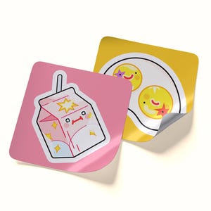 May include: Two square stickers with rounded corners. One sticker is pink and features a white milk carton with a smiling face and yellow star accents. The second sticker is yellow and displays a white plate with two fried eggs with faces.