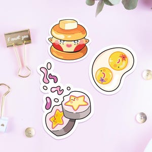 May include: Three die-cut stickers featuring cartoon food characters: a pancake stack with a butter pat, a fried egg with two yolks, and two sushi rolls with star-shaped centers. Two metal binder clips and small metal studs are also present.