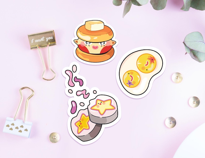 May include: Three die-cut stickers featuring cartoon food characters: a pancake stack with a butter pat, a fried egg with two yolks, and two sushi rolls with star-shaped centers. Two metal binder clips and small metal studs are also present.