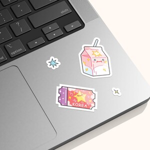 May include: A collection of four vinyl stickers featuring a pink milk carton with a straw, a purple and pink ticket with a yellow star and the text KOREA, a blue four-pointed star, and a small white four-pointed star, all applied to a laptop.