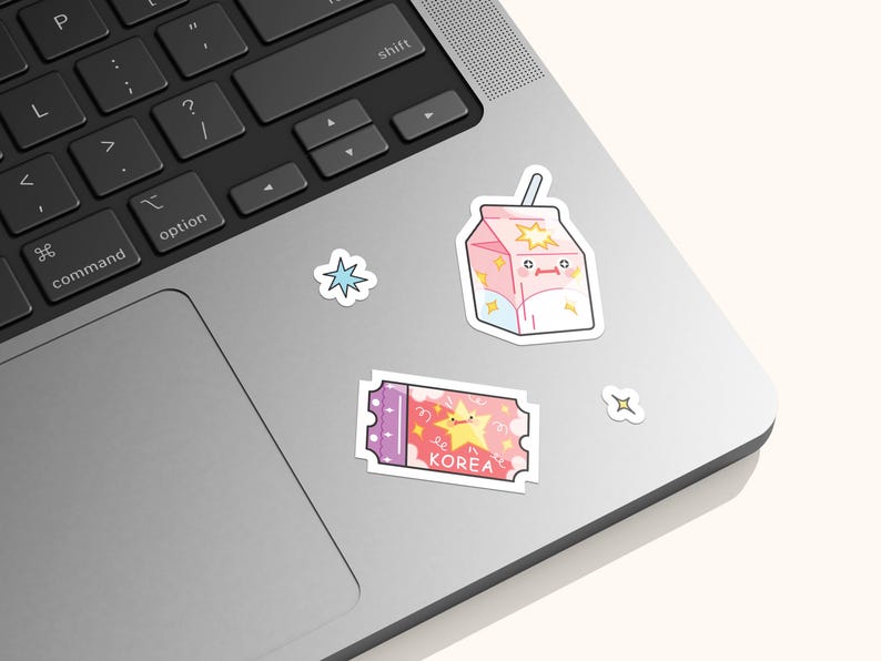 May include: A collection of four vinyl stickers featuring a pink milk carton with a straw, a purple and pink ticket with a yellow star and the text KOREA, a blue four-pointed star, and a small white four-pointed star, all applied to a laptop.