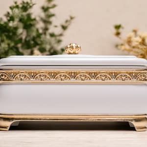 May include: A white decorative box with gold accents. The box has a curved shape with a gold filigree design around the middle and base. A gold knob sits atop the lid. The box is on a wooden surface.