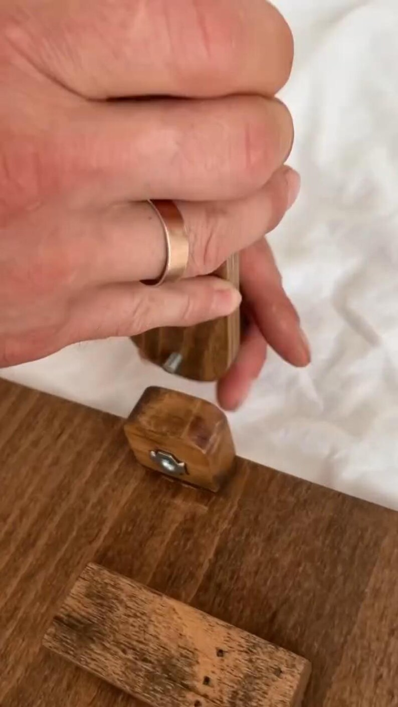 May include: Wooden furniture assembly in progress. A person is using a tool to connect two wooden pieces. The pieces are a warm brown color, and the tool is also wooden. The background is a soft, white fabric.