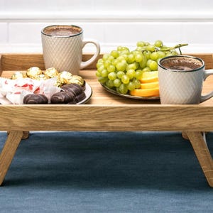 May include: A wooden breakfast tray with folding legs holds two mugs of coffee, a plate of chocolates and wrapped candies, and a plate of green grapes and orange slices. The tray is set on a blue textile.
