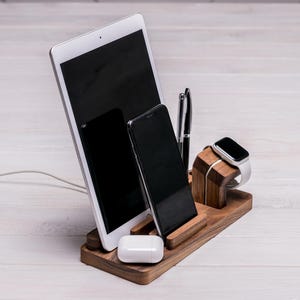 以下が含まれることがあります： 木製のデスクオーガナイザーには、タブレット、スマートフォン、ワイヤレスイヤホンが収納されています。ペンとスマートウォッチもオーガナイザーに入っています。オーガナイザーは濃い色の木材でできており、白い木製の表面に置かれています。