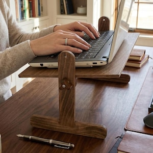 May include: A wooden laptop stand with a dark brown finish. The stand is adjustable and has a T-shaped base. A person is typing on a laptop placed on the stand. A pen rests on the desk in front of the stand.