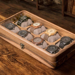 May include: A wooden watch display case with a hinged lid, containing nine watches. The watches feature diverse strap and face designs, including black, brown, and wood tones. The box is made of light-coloured wood and rests on a wooden surface.