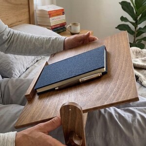May include: A wooden bed desk with a book and pen on top. The desk is made of light brown wood and has a slanted surface. The book is dark blue. The desk is supported by a wooden stand.
