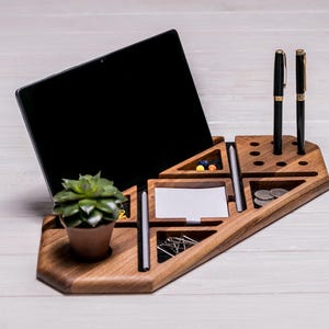 Könnte beinhalten: Schreibtisch-Organizer aus Holz mit mehreren Fächern. Er verfügt über einen Steckplatz für ein Tablet, Stifthalter, eine kleine Topfpflanze und Platz für Papier und andere Gegenstände. Der Organizer ist aus hellbraunem Holz gefertigt, ca. 30 cm lang.