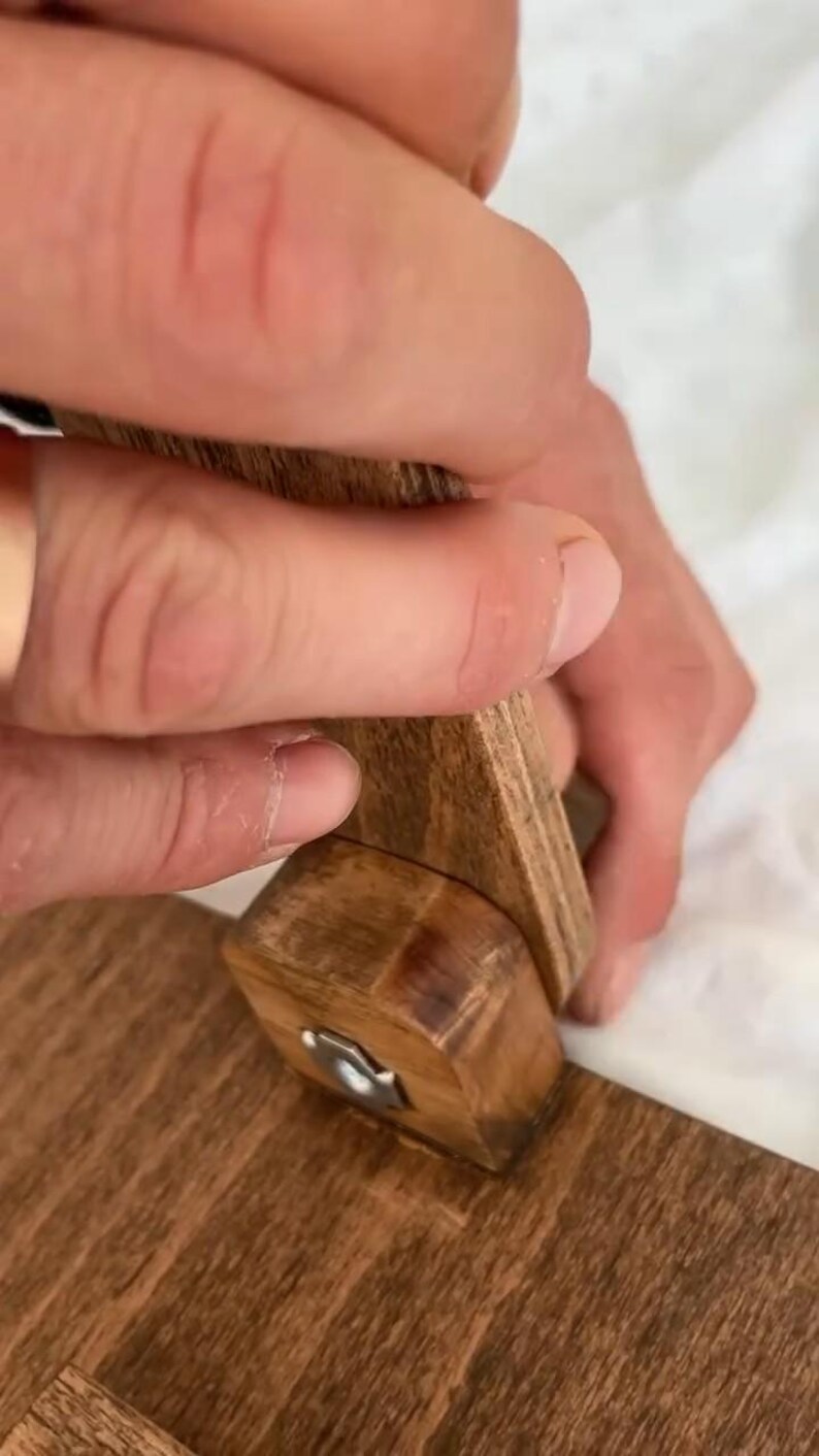 May include: Close-up of a wooden object being assembled. The object is made of light brown wood and features a metal hinge. The hands are holding the wooden pieces together, suggesting a construction or repair process.