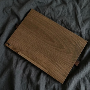 May include: A rectangular wooden book stand with a rich brown finish. The stand features a small ledge to hold pages and a support piece. The wood grain is visible. The word "REMEDIO" is printed on the stand.