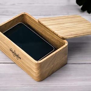 May include: A wooden box with a partially open lid, showing a black smartphone within. The box is made of light-coloured wood with a visible grain. The text "WOODEN" is printed on the side.