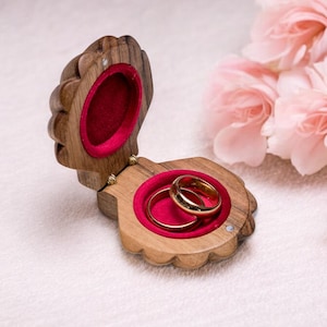 Puede incluir: Un joyero de madera con forma de concha, abierto para revelar dos alianzas de oro en el interior de terciopelo rojo. La caja se coloca sobre un fondo de suaves flores rosas, creando una escena romántica.