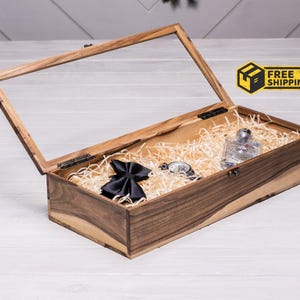May include: A wooden gift box with a clear glass lid, containing a black bow tie, a silver watch, and a clear glass bottle. The box is filled with straw-like material. The box is open, and the lid is propped up.