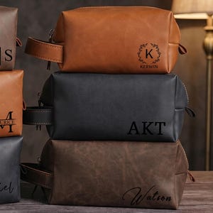 Op de afbeelding: Een stapel gepersonaliseerde leren toilettassen in bruine en zwarte tinten. Elke tas heeft een monogram of naam in een contrasterende kleur. De tassen hebben een ritssluiting en een handvat. Afmetingen in centimeters.