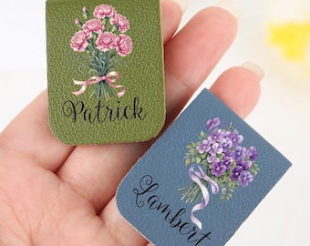 May include: Two leather bookmarks are held in a hand. One is olive green with a pink flower bouquet and the name "Patrick". The other is blue with a purple flower bouquet and the name "Lambert".
