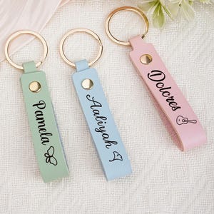 May include: Four personalized keychains in olive green, mint, light blue, and pink leather. Each keychain has a gold ring and a name printed in black script. The pink keychain has a guitar graphic.