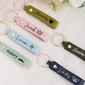 May include: A collection of personalized keychains in various colors, including green, pink, blue, and navy. Each keychain features a gold ring and is engraved with a name and a small icon, such as a heart, crown, or butterfly.