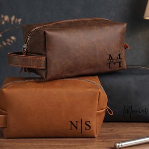 May include: Three leather toiletry bags in brown, tan, and black, each with a zipper closure and a handle. The bags are personalized with initials or names. The brown bag has the initials "M" and "T", the tan bag has "N" and "S", and the black bag has the name "Muriel".