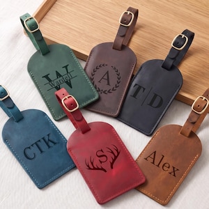 May include: Six rectangular leather luggage tags in green, brown, dark gray, blue, red, and tan. Each tag features a gold-toned metal buckle on a strap and is personalized with unique laser-engraved monograms, names, or decorative motifs.