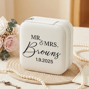Puede incluir: Una caja de joyería cuadrada blanca con cierre de cremallera. La caja está inscrita con "MR. & MRS. Browns 1.9.2025" en escritura negra. La imagen también incluye un ramo de rosas, un collar de perlas y un frasco de perfume.