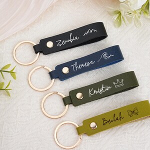 May include: Four personalized leather keychains in black, navy, olive green, and yellow-green. Each keychain has a gold-colored ring and a name or word in white script. The black keychain has a mountain design, the navy has a wave, the olive green has a crown, and the yellow-green has a bow.