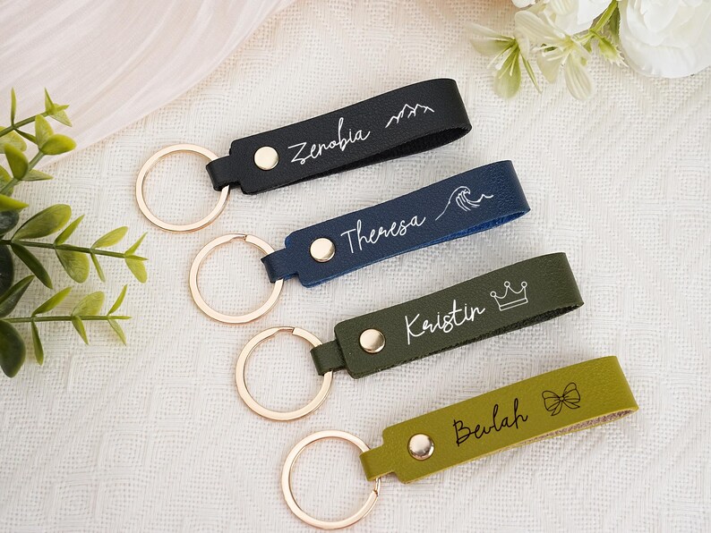 May include: Four personalized leather keychains in black, navy, olive green, and yellow-green. Each keychain has a gold-colored ring and a name or word in white script. The black keychain has a mountain design, the navy has a wave, the olive green has a crown, and the yellow-green has a bow.