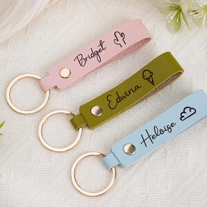 May include: Three personalized keychains in pink, olive green, and light blue. Each keychain features a name and a small icon: a cactus, an ice cream cone, and a cloud. The keychains have gold-colored key rings.