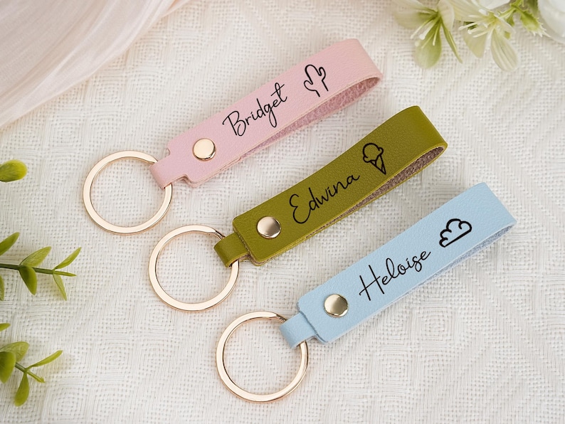 May include: Three personalized keychains in pink, olive green, and light blue. Each keychain features a name and a small icon: a cactus, an ice cream cone, and a cloud. The keychains have gold-colored key rings.