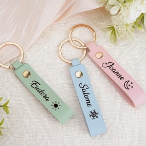 May include: Three personalized keychains in pastel colors: mint green, light blue, and pink. Each keychain has a gold ring and a leather strap with a name and a small graphic. The mint green keychain has the name "Eudora" and a sun graphic.