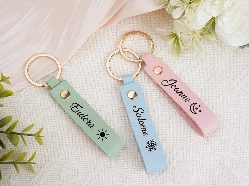 May include: Three personalized keychains in pastel colors: mint green, light blue, and pink. Each keychain has a gold ring and a leather strap with a name and a small graphic. The mint green keychain has the name "Eudora" and a sun graphic.