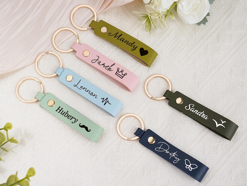May include: A collection of personalized keychains in various colors, including green, pink, blue, and navy. Each keychain features a gold ring and is engraved with a name and a small icon, such as a heart, crown, or butterfly.