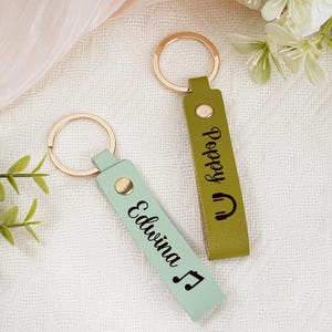May include: Two personalized keychains with gold-colored rings. One is light green with the name "Edwina" and a musical note. The other is olive green with the name "Poppy" and a headphone icon. Both have a gold snap.