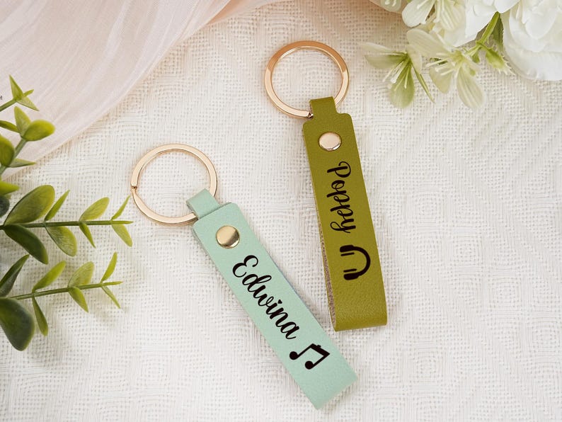 May include: Two personalized keychains with gold-colored rings. One is light green with the name "Edwina" and a musical note. The other is olive green with the name "Poppy" and a headphone icon. Both have a gold snap.
