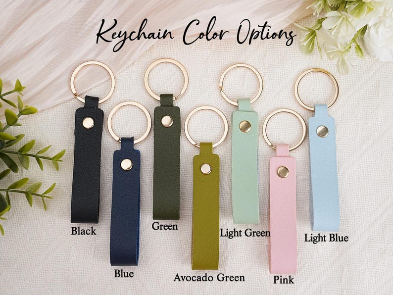 May include: A selection of leather keychains in various colors, including black, blue, green, avocado green, light green, pink, and light blue. Each keychain has a gold-colored ring and a matching snap closure. The text "Keychain Color Options" is at the top.